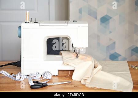 Sewing machine with piece of beige fabric, scissors, measuring tape and pins on table, tailor workplace. Stock Photo