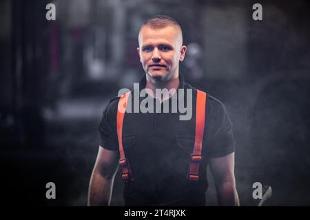 firefighter portrait wearing shirt and red throuser suspenders. holding an axe. smoke and fire trucks in the background. Stock Photo