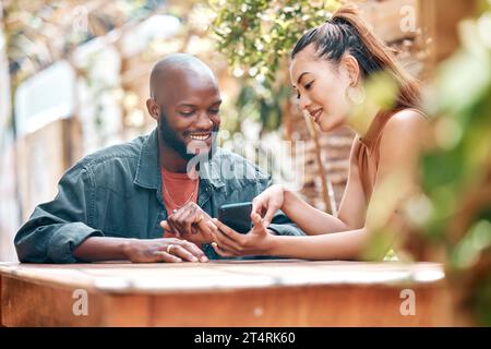 Interracial couple using a phone together while sitting outside at a cafe. Mixed race woman showing an african american man a message on her phone Stock Photo