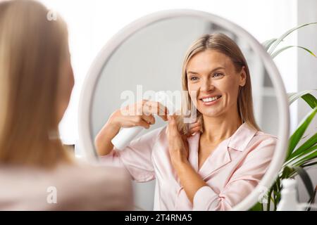 Haircare. Smiling Middle Aged Woman Applying Hair Spray While Standing Near Mirror Stock Photo