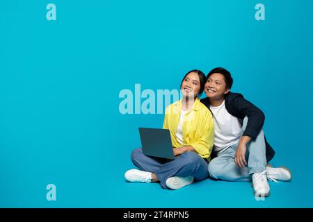 Smiling millennial asian couple in casual use laptop, sit on floor, look at free space Stock Photo