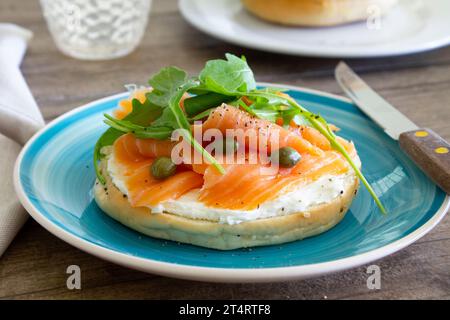 Smoked salmon bagel with cream cheese, capers and arugula served on a light blue plate on a rustic wooden table. Stock Photo