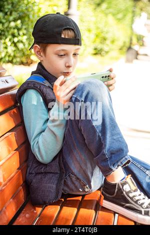 Pre-adolescent boy sitting on a park bench in autumn looking at his mobile phone, Belarus Stock Photo