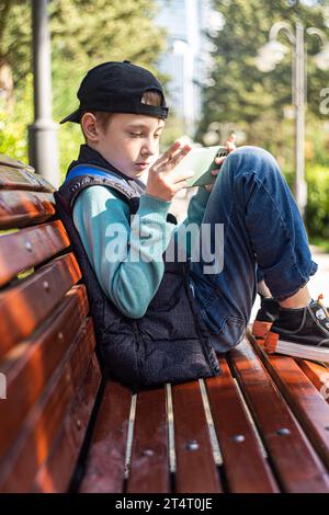 Pre-adolescent boy sitting on a park bench in autumn looking at his mobile phone, Belarus Stock Photo