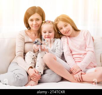 Portrait of mom hugging her cute daughters while sitting on the sofa and watching TV at home. Happy family relaxing on the coach Stock Photo