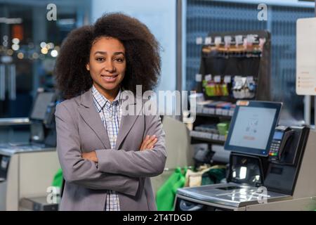 Portrait female manager of a supermarket store, a business owner with folded hands smiling and looking at the camera, a businesswoman on the background of self-service checkouts in a business suit. Stock Photo