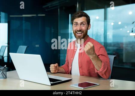 Portrait of successful mature businessman in red shirt at workplace inside office, man celebrating victory and successful achievement results at work, looking at camera, holding hand up. Stock Photo