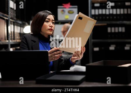 Asian private detective in agency file cabinet room reading criminal case file details. Investigator woman surrounded by criminology document folders on office repository cabinet shelves Stock Photo