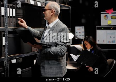 Senior private investigator in agency depository office room looking for criminal case file paperwork on cabinet shelves. Elderly detective and asian assistant surrounded by criminology folders Stock Photo