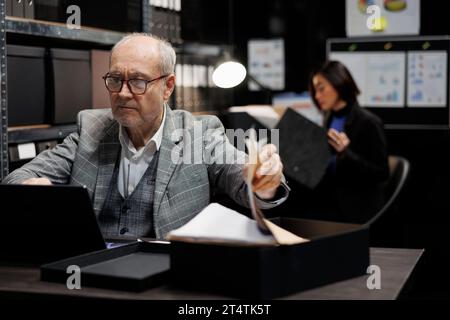 Elderly private investigator flicking through criminal case details paperwork before starting investigation. Senior detective in archive room office filled with criminology folder files Stock Photo