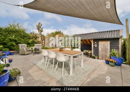 a patio area with chairs, tables and an umbrella over the dining table in front of a wooden shed behind Stock Photo