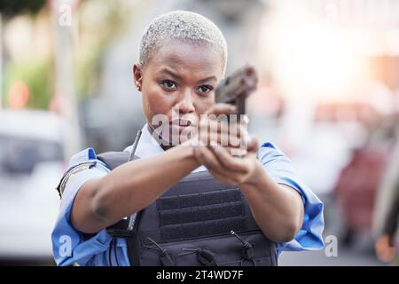 Gun, serious and female police officer in the city for an arrest to stop crime in a street. Upset, young and African woman security guard with pistol Stock Photo