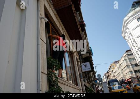 Woman in red effortlessly dangles outside a hotel lobby window, embracing the daring task of cleaning it amidst the urban backdrop. Stock Photo