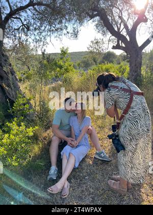 Photographer leaning over to take pictures of man and woman embracing, sitting on the grass Stock Photo