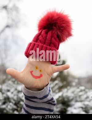 smiling face is drawn on the child palm, and a warm red knitted hat is put on his fingers. It's time to warm up and enjoy winter walks. happy childhoo Stock Photo