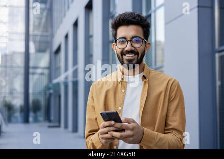 Portrait of young successful Indian businessman, man smiling and looking at camera from outside office building, programmer in shirt holding phone, typing message, and using app. Stock Photo
