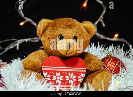 A brown teddy bear sits with a Christmas tree toy in the form of a heart on white tinsel, surrounded by red Christmas tree decorations. In the backgro Stock Photo