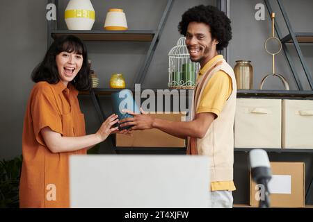 Young cheerful intercultural couple showing vase from new collection and looking at laptop screen while communicating with online audience Stock Photo