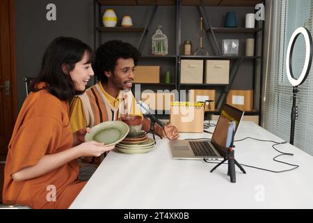 Two happy young bloggers with unpacked handmade earthenware sitting in front of laptop during livestream and speaking to online audience Stock Photo