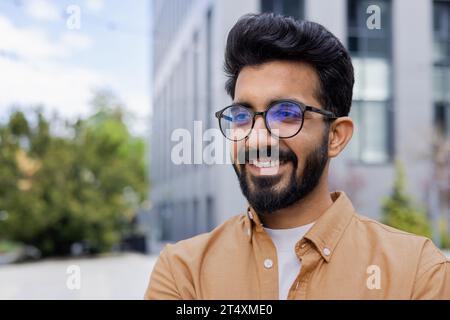 Young joyful man close-up smiling and looking away, Indian man in glasses outside office building walking in the city, programmer student in shirt. Stock Photo