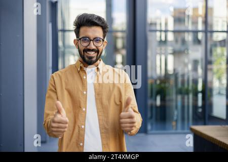 Portrait of a young successful Indian businessman, man smiling and looking at the camera from outside an office building, showing the camera a thumbs up affirmatively and recommending. Stock Photo