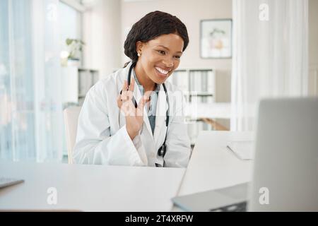 Video call, virtual medical meeting with a doctor waving, greeting and smiling with a laptop online working at a hospital. Professional healthcare Stock Photo