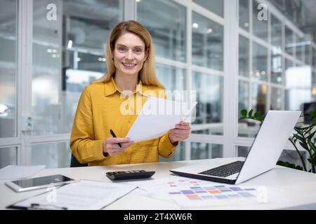 Portrait happy successful woman financier doing paperwork, businesswoman holding papers, documents, contracts and reports in hand, smiling joyfully, satisfied working with laptop, using calculator Stock Photo