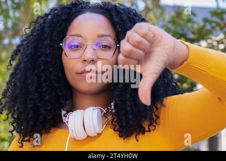 portrait Young angry african american woman pointing thumb down looking thoughtful outdoors.  Stock Photo