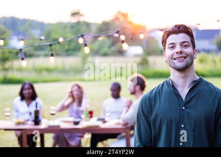 An attractive South European man with short brown hair in a green shirt laughs joyously with a wine glass in hand, as friends share the mirth in a garden barbecue setting, under the warm sunny skies. The scene is a lively blend of camaraderie, good food, and the gentle embrace of nature, epitomizing a perfect outdoor picnic. Charming Young Man Relishing Sunny BBQ Gathering with Friends. High quality photo Stock Photo