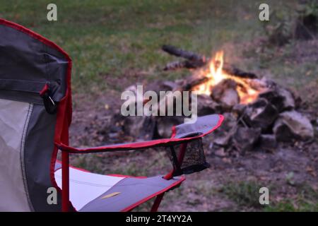 A camp chair sitting in front of a campfire, focus on chair in foreground, background blurred.  A time of relaxation and getting back to nature. Stock Photo