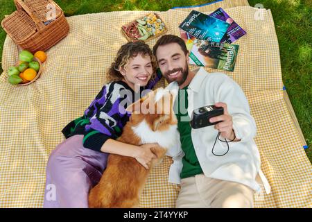 top view of happy couple taking photo with cute corgi dog on camera while having picnic in park Stock Photo
