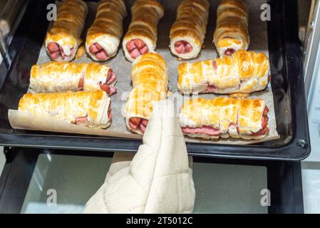 Pulling out a baking sheet with baked sausages in dough from the oven. The process of preparing sausages in dough at home. Stock Photo