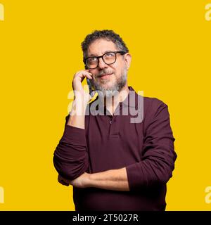 A thoughtful middle-aged man with glasses and a gray beard engages in a phone conversation, wearing a maroon shirt against a vibrant yellow backdrop, Stock Photo