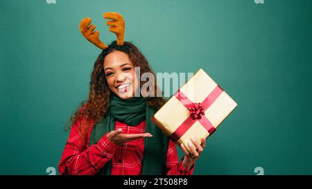 Excited young woman gestures towards Christmas gift, palm up, studio shot Stock Photo