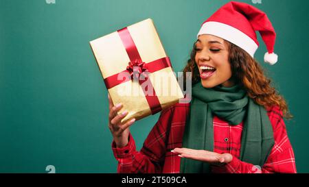 Excited young woman gestures towards Christmas gift, palm up, studio shot Stock Photo