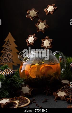 Glass teapot with brewed green tea with citrus fruits among fir branches and Christmas decorations, flying cookie and spices Stock Photo