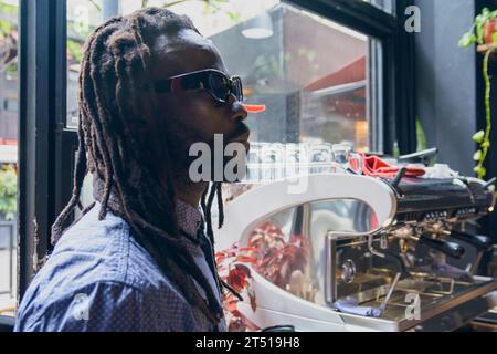 young man of African ethnicity with dreadlocks, beard and glasses, calm standing at restaurant counter looking at menu on wall and thinking what to or Stock Photo
