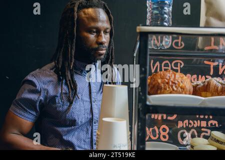 young African man with dreadlocks and beard, hungry standing inside restaurant looking at candy counter deciding what he wants to order to eat Stock Photo
