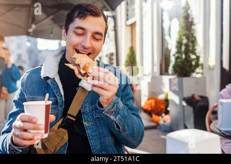 Young happy man having breakfast with coffee and croissant sitting at outdoor cafe in European city. Tourist enjoys street food. Stock Photo