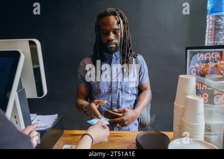 young black man with dreadlocks and beard standing in front of restaurant counter receiving his cards from waiter after paying for food order Stock Photo