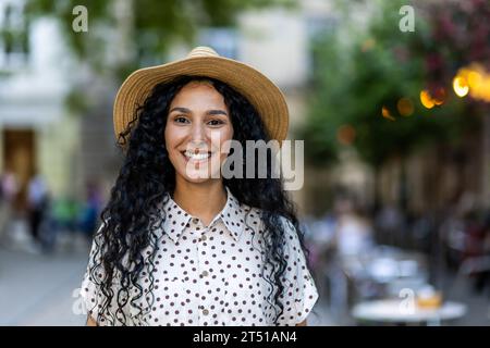 Beautiful young Latin American woman portrait, woman walking in evening city in hat with curly hair in warm weather, smiling and looking at camera close up. Stock Photo