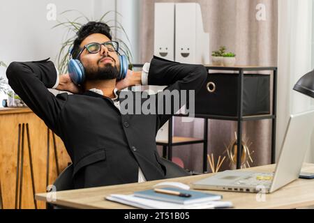 Happy smiling Indian businessman listening music on headphones taking break relaxing leaning on chair after working at office workplace. Male Hindu freelancer in formal suit puts hands behind head Stock Photo