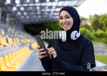 Portrait of a young hijab Muslim woman in a sports stadium running and doing active physical exercises, smiling and looking at the camera, using the phone and headphones to listen to music. Stock Photo