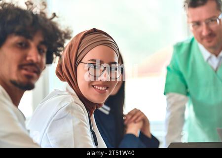 A smiling female doctor in a hijab participates in a positive and collaborative meeting with her colleagues, exemplifying diversity and teamwork in a Stock Photo