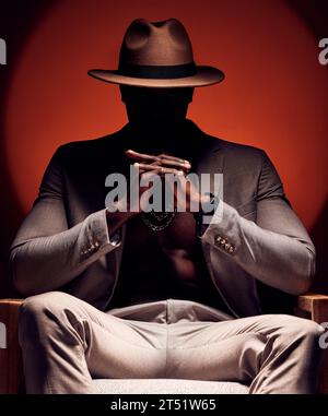 Stylish, fashionable and trendy man in silhouette sitting in a dark room with a red studio background. Edgy, mysterious and curious male mafia boss Stock Photo