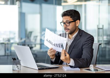 Serious successful businessman inside office on online video meeting presentation, man in business suit shows report graph to camera, uses laptop for video call at workplace. Stock Photo