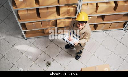 Female supervisor checking list of products on racks, looking at cardboard boxes to plan distribution. Woman with hardhat working in warehouse with merchandise and stock, storage inventory. Stock Photo