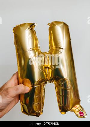 A male hand showcases the letter H, crafted from gold helium balloons. It gleams prominently against a pure white background, perfect for special occasions. Stock Photo