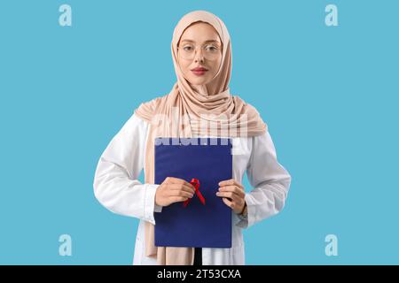 Female Muslim doctor with red ribbon and clipboard on blue background. World AIDS Day concept Stock Photo