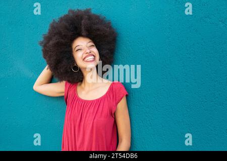 Portrait of smiling Moroccan woman in casual clothes with Afro hairstyle standing against bright blue wall Stock Photo